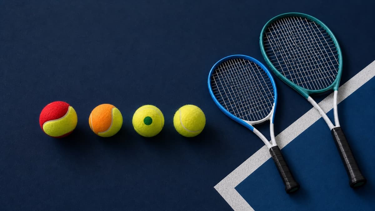Red-and-yellow, orange-and-yellow, green-dot and standard yellow tennis balls beside two junior rackets.